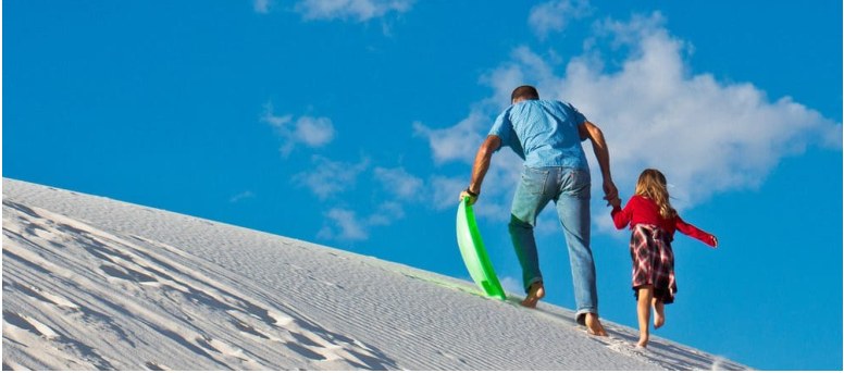 An adult and child walking up a white sand dune with a sled.